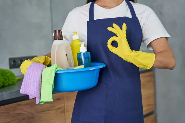 Best products. Cropped shot of cleaning lady showing ok sign while holding container full of cleaning products and equipment, standing in the modern kitchen