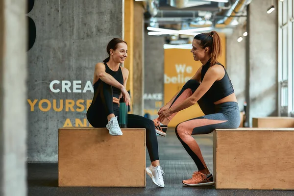 Full length of two young athletic fitness girls in sportswear sitting on crossfit jump boxes at gym and talking, exercising together