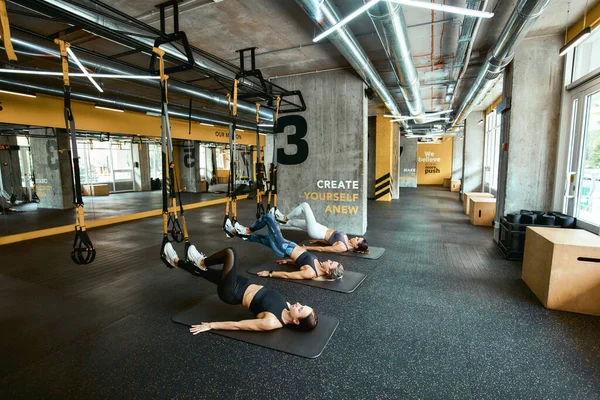 Strength training. Three young caucasian athletic women in sportswear exercising with fitness trx straps at industrial or crossfit gym