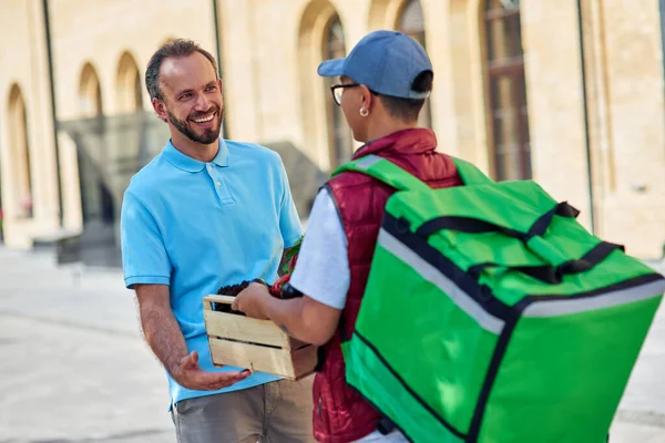 Cheerful caucasian man customer receiving wooden box with fresh grocery from male courier with thermo bag while standing outdoors on the sunny street