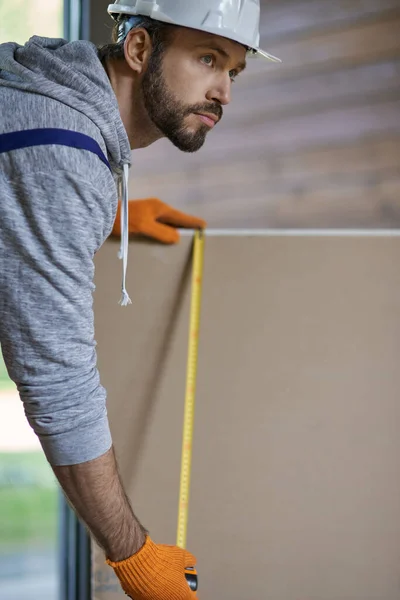 Close up shot of confident young male builder in hard hat measuring drywall while working at construction site — Stock Image Close up shot of confident young male builder in hard hat measuring drywall while working at construction site Stock Photo