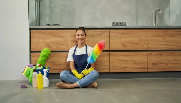 Safe and clean. Joyful young woman smiling at camera, holding cleaning duster while resting after cleaning apartment with detergents