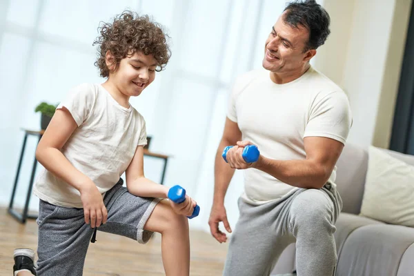 Family workout. Sportive latin middle aged father teaching his son exercising with dumbbells while spending time together at home