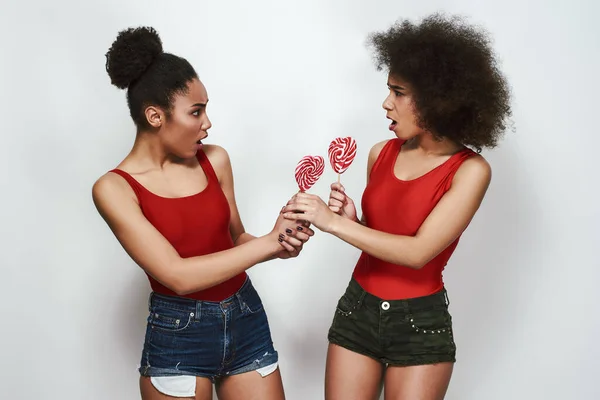 I want more Two angry afro american woman in jeans shorts are looking at each other with opened mouth and holding lollipops shaped heart