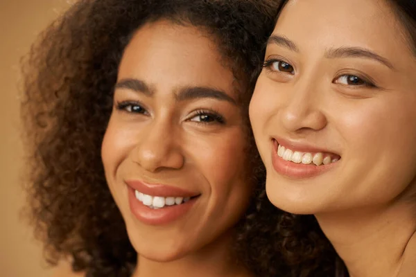 Face closeup of two joyful attractive mixed race young women with perfect smile posing for camera isolated over beige background