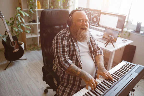 Joyful obese man artist plays music on synthesizer near table with computer monitors in studio