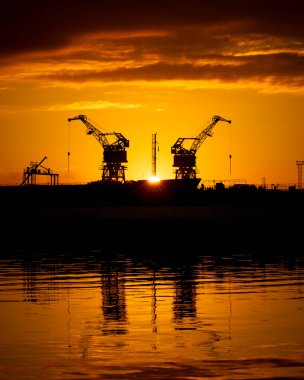 Limandaki batan güneş diskine karşı güzel metal vinç siluetleri. Riga, Letonya 'nın sanayi şehri.