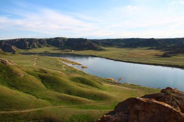 Güneşli bir günde bozkırda nehir. Sakin bir nehir, bozkır, alçak dağlar, tepeler, açık mavi bir gökyüzü nehirde yansıyor, yeşil çayırlar nehrin etrafında, ön planda büyük sarı taşlar var.