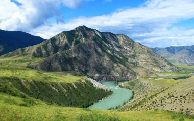 The Katun River in Altai in a high-mountain valley, in the background there are high rocky mountains of the ridge, near the river there are high banks, summer, sunny
