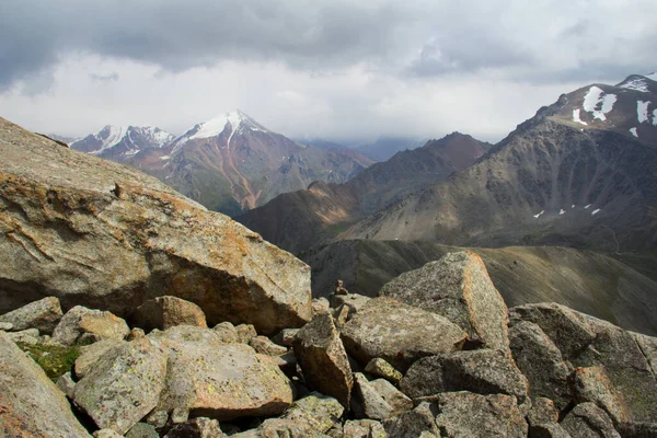 A large pile of huge stones in the foreground, in the distance the snow-capped peaks of the ridges against the background of a gloomy sky with clouds, Big Almaty peak, summer, sunny