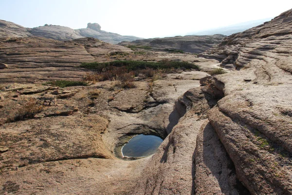 Volcanic layered mountains in the Bektau-Ata tract rise in layers, in front of a small puddle of water, green low bushes, slopes in the distance, summer, sunny
