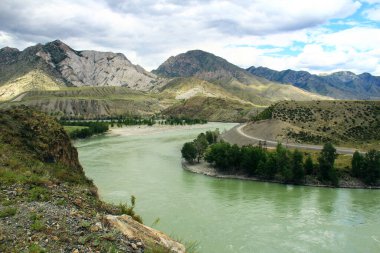 Altai 'deki yüksek dağlık bir vadide, Katun Nehri akıyor, uzaklarda dağlar var, kıyıda ağaçlar yetişiyor, yamaç boyunca bir yol, alçak bulutlu bir gökyüzü, yaz, güneşli