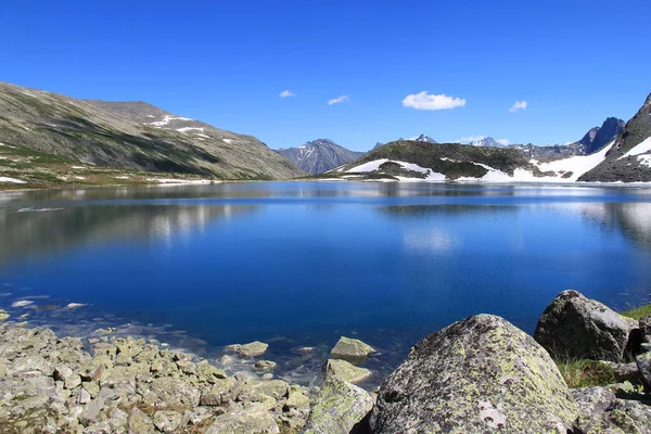 Altai 'deki yüksek dağ gölünün aynası, dağlarla çevrili tepeler ve karla kaplı tepeler, gölün kıyısındaki taşlar, bulutlu gökyüzü, yaz, güneşli