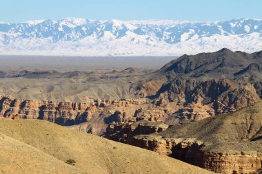 Yazın karla kaplı yüksek dağ sırtı olan uzak bir vadi manzaralı Charyn Kanyonu, güzel bir rahatlama deseni olan kanyon duvarları, yaz, güneşli