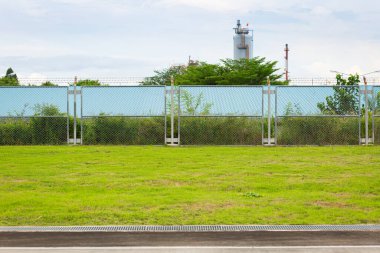 Ön planda yeşil çimenli çit, güvenlik, sınır, açık hava manzarası ve organize endüstriyel ya da yerleşim ortamını temsil ediyor..