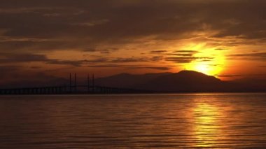 Su, turuncu gündoğumunda denizde yavaşça akar. Arka plan Penang Köprüsü ve tepe..