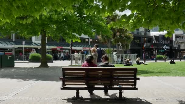 Un couple s'assoit sur le banc le matin brillant. La promenade touristique au bord du lac dans le centre de Queenstown. — Video
