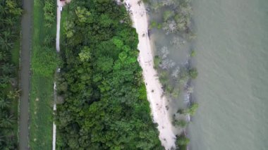 Kumsalın yanında yeşil bir orman var. Kumsal kumlu ve birkaç ağacı var. Su sakin ve suda kayıklar var. Hava görüntüsü Pantai Bukit Batu