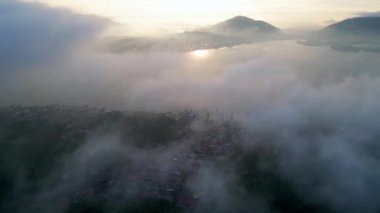 Gökyüzü sisle kaplı ve güneş Pulau Pangkor 'da batıyor. Dağlar arka planda ve su sakin.