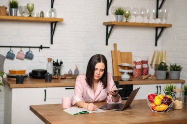 Uzaktan öğrenme çevrimiçi. Sınavlar için hazırlık. Elinde telefon olan bir kadın bilgisayarın başına oturmuş bir şeyler yazıyor. Uzak çalışma.