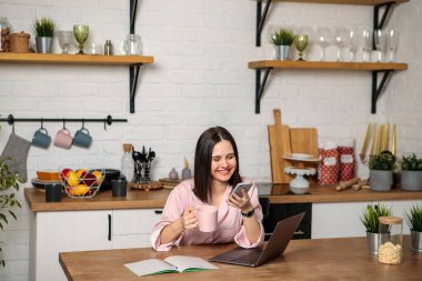 Uzaktan kumanda. Mutfakta bilgisayarın başında oturan bir kadın telefonla konuşuyor. Çevrimiçi uzaklık öğrenimi, eğitim ve çalışma.
