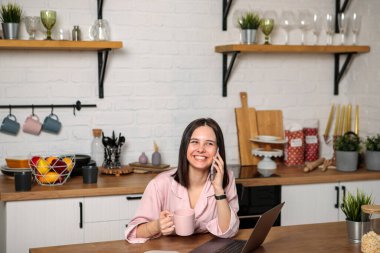 Uzaktan kumanda. Mutfakta bilgisayarın başında oturan bir kadın telefonla konuşuyor. Çevrimiçi uzaklık öğrenimi, eğitim ve çalışma.