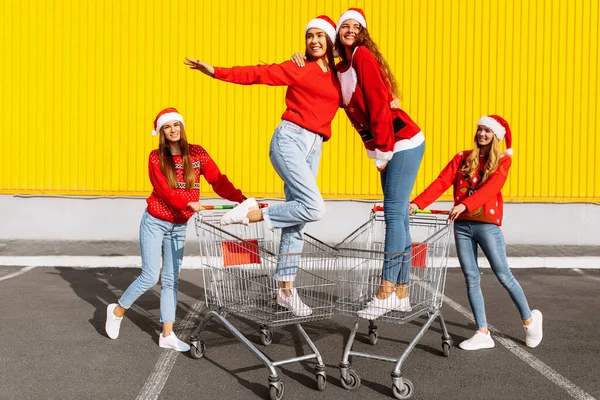 Group of four young women in Christmas sweaters and Santa Claus hats having fun on carts near a shopping center