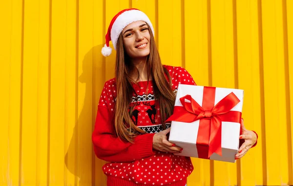 Attractive happy young woman in christmas sweater and santa claus hat with christmas present outdoors