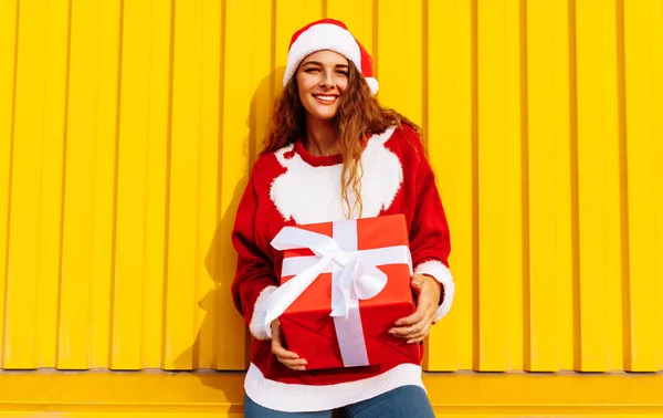 Attractive happy young woman in christmas sweater and santa claus hat with christmas present outdoors