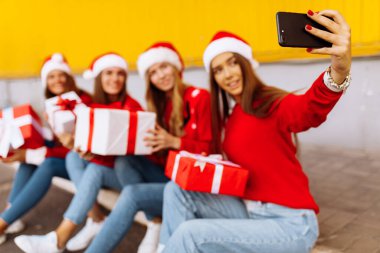 Noel Baba şapkalı bir grup mutlu genç kadın ellerinde hediye kutuları ve cep telefonlarıyla dışarıda sarı duvar arkasındayken selfie çekiyorlar.