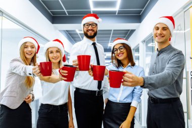 Noel Baba şapkalı başarılı genç işadamları bayramı kutluyor ve modern bir ofiste kırmızı bardaklardan bir içki içiyorlar. Mutlu yıllar ve mutlu Noeller..