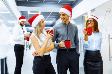 Noel Baba şapkası takan bir grup mutlu iş adamı ofiste yeni yılı kutlarken konuşuyor ve gülümsüyor. Yeni Yıl ve Mutlu Noeller