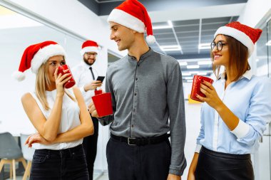 Noel Baba şapkası takan bir grup mutlu iş adamı ofiste yeni yılı kutlarken konuşuyor ve gülümsüyor. Mutlu yıllar ve mutlu Noeller.