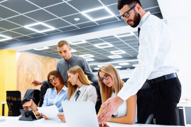 Yeni bir proje üzerinde çalışan iş ekibi modern ofislerinde masalarında otururken gülümsüyor.