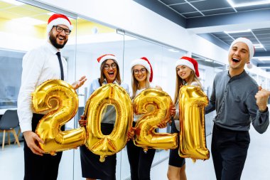 Noel Baba şapkalı bir grup neşeli iş arkadaşı, eğleniyor ve yeni yılı kutluyorlar, 2021 'in bir sembolü olan altın rakamlı balonlar tutuyorlar.