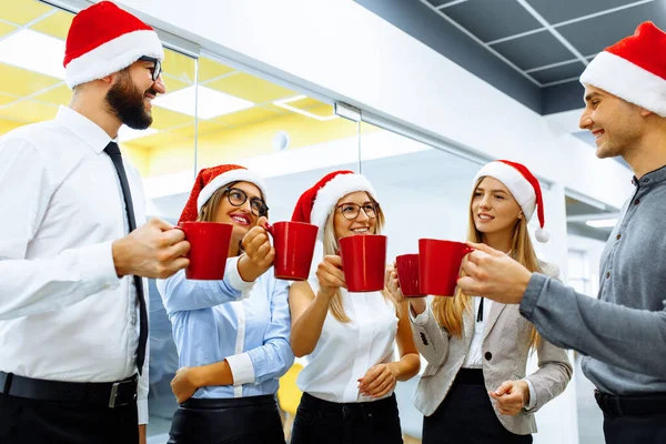 group of business people in Santa Claus hats, Colleagues celebrate New Year in the office, have fun and drink drinks from red mugs