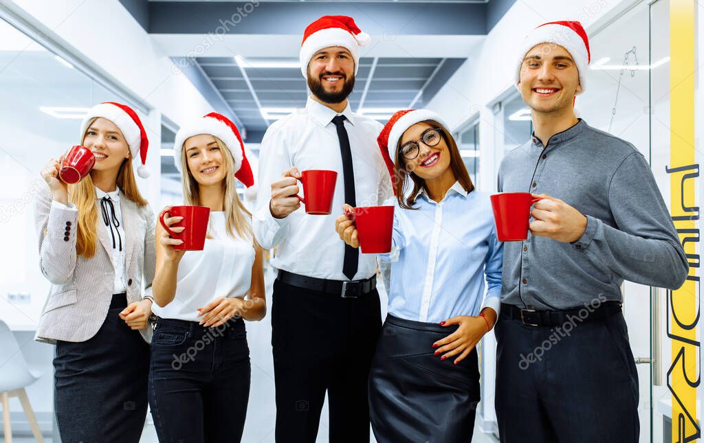 Successful young businessmen in Santa Claus hats celebrate the holiday and drink a drink from red mugs in a modern office. Happy New Year and Merry Christmas.