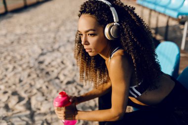Genç atletik Afrikalı Amerikalı kadın. Kulaklık takıyor. Yoğun bir koşudan sonra dinleniyor ve dışarıda koştuktan sonra şişeden su içiyor.