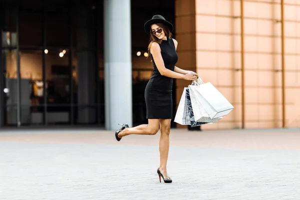 黒のドレスと帽子の陽気なスタイリッシュな幸せな女の子は 買い物に行くのが大好き 彼女はショッピングバッグを運び 最新のオファーのためにモールに走ります ショッピング ブラック フライデー — ストック写真