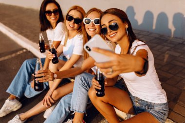 Cam şişelerde içen mutlu gençler, birlikte vakit geçirir, cep telefonuyla selfie çekerler, güneşli bir yaz gününde, sarı bir duvarın arkasında.