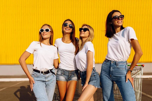 サングラスや夏の服を着た幸せな若い女性 市内の黄色の壁の背景にカートで楽しんでいる肯定的な友人 — ストック写真