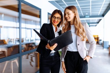İş kadınları ofiste koridorda yürürken belgeleri tartışıyor.