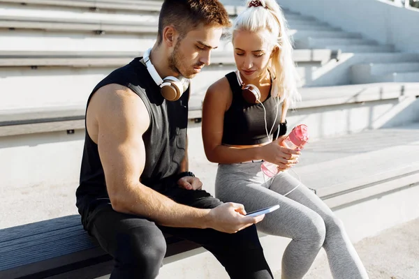 Young sports couple, in sportswear, sitting on the stairs outdoors, listening to music on headphones and using a mobile phone, fitness people after a successful workout