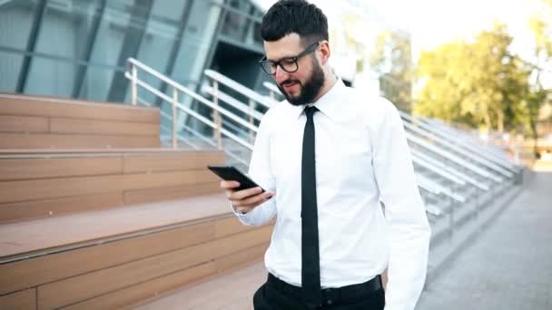 Hombre de negocios con barba con gafas, vestido con ropa elegante, usando un teléfono móvil, un joven hombre de negocios leyendo algo en un teléfono móvil durante un descanso al aire libre — Vídeos de Stock