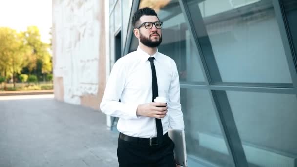 Glücklicher junger Geschäftsmann hält eine Tasse Kaffee in der Hand, während er draußen vor dem Hintergrund des Business Centers spaziert — Stockvideo