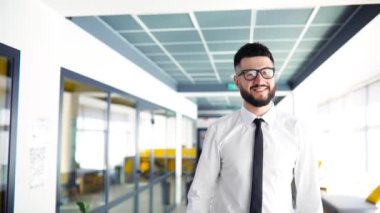 İş kıyafetleri içinde sakallı ve gözlüklü bir işadamı modern bir ofisin koridorunda yürüyor.