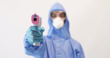 Erkek doktor koruyucu giysi, gözlük, maske ve eldiven takıyor, sıcaklığı ölçmek için temas olmayan bir termometreye sahip, beyaz arka planda, koronavirüs salgını 