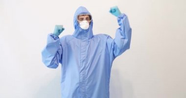 Covid-19 'la dövüşmek için koruyucu giysili bir doktor, gözlük, maske ve eldiven takıyor, beyaz bir arkaplanda zafer jesti gösteriyor.