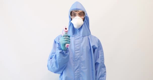 Médico masculino vestindo roupas de proteção, óculos, máscara e luvas, com um termômetro sem contato para medir a temperatura, em um fundo branco, epidemia de coronavírus — Vídeo de Stock