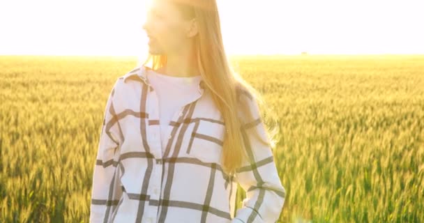 Uma mulher bonita em uma camisa verificada caminha através de um campo de trigo ao pôr do sol — Vídeo de Stock